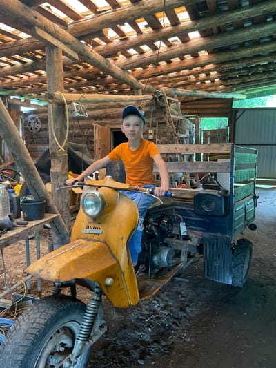 A young boy wearing an orange t-shirt and a blue baseball cap sits on a weathered, yellow three-wheeled motor vehicle with a blue cargo bed in a rustic, wooden structure that appears to be a shed or workshop. The structure has a roof made of logs and planks, allowing sunlight to filter through. The motor vehicle has a scooter-like front with a large headlight and a prominent yellow fender. The boy's hands are on the handlebars. Behind him, the cargo bed is loaded with items, and a black jerry can is attached to the side. Various tools and objects, including bottles and a bucket, are visible on a workbench to the left of the vehicle. The floor is dirt and scattered with debris. The environment suggests a rural or semi-rural setting.