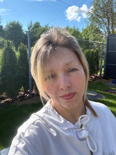 A woman with blonde hair is taking a selfie outdoors. She is wearing a white hoodie with a drawstring closure. Her left eye is visible, as is her mouth, which appears to be slightly open. The background is a blurred outdoor scene with green grass, trees, a fence, and a bright blue sky with white clouds. The lighting suggests it is daytime, likely sunny. The image is oriented vertically, with the woman's head tilted to the right. There is no visible text in the image. The location context provided is Жостово, Russia.