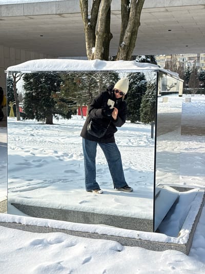 The scene depicts a snow-covered urban park in Plodorodny, Russia, on a bright winter day. A rectangular art installation featuring mirrored sides rests on a grey granite base, both partially covered with snow. A female individual is captured in the mirror's reflection, positioned centrally. She is wearing a cream knit beanie, dark sunglasses, a black puffer jacket, black gloves, blue denim jeans, and athletic shoes (one light-colored, one dark). She holds a white smartphone horizontally with both hands, angled as if recording or photographing herself. A black crossbody bag is worn over her left shoulder. The background, visible in the mirror's reflection and directly beyond the structure, consists of snow-dusted bare deciduous trees, dark evergreen trees, and distant multi-story residential buildings. A yellow illuminated angelic figure and a red building are also visible in the reflected background to the left. The ground surrounding the installation is uniformly covered in snow, with footprints visible on the granite base.