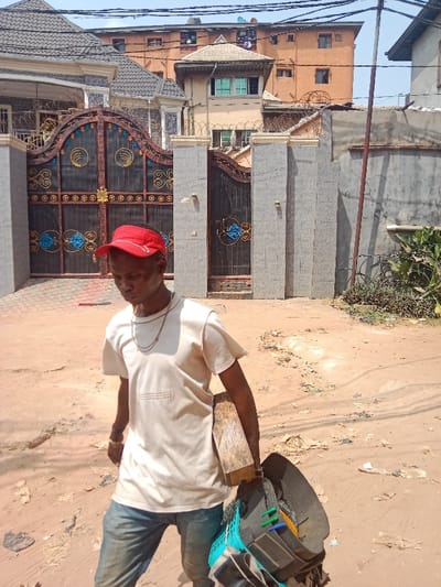 A male individual is observed walking on an unpaved, reddish-brown dirt road in Awada, Nigeria. He is wearing a red visor, a light-colored short-sleeved t-shirt with embroidered detailing on the chest, blue jeans, and a chain necklace. He carries a rectangular wooden block under his right arm and a dark bag with light blue mesh and assorted items in his left hand. His gaze is directed downwards and slightly forward.

The immediate background features a large, ornate dark metal gate with gold and blue decorative elements, set into light grey pillars topped with barbed wire. Behind the gate, a multi-story residential building with a dark grey tiled roof and a smaller annex building with green-tinted windows are visible. To the right of the gate structure, a grey wall and a concrete fence with cracks are present, alongside green foliage. A dark utility pole with overhead wires stands on the far right. The distant background reveals a multi-story building with light brown walls and multiple windows, under a clear, bright sky.