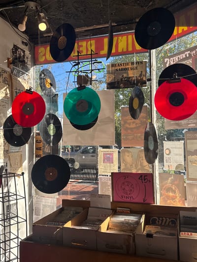 A record store window display showcases colorful vinyl records—red, green, and black—suspended on thin strings. Posters and album covers adorn the glass, including one featuring the Beastie Boys' "Sabotage." Inside, shelves packed with records are organized alphabetically and by genre (Reggae). A sign indicates 45 rpm records are available. The reflections reveal an urban street scene with buildings and passing cars. The display mixes vintage charm with modern music culture.