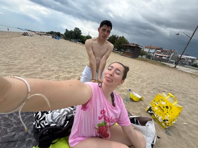 A selfie, taken by a female, shows her and a male companion on a beach at Νέα Φλογητά, Greece. The female, positioned at the top of the frame, wears a pink t-shirt. The male, below her, is shirtless. The beach setting includes sand, buildings, trees, and cloudy skies. A yellow bag and a bottle are among the items on the sand near the female.