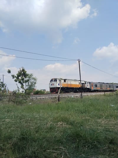 The image depicts a train moving along a railway track in Gebang, Indonesia. The train consists of two locomotives and passenger cars. The lead locomotive has a white and orange color scheme with the logo "KAI" visible on the side. The second locomotive and passenger cars appear to be gray and orange. Overhead power lines are present, supported by utility poles. The foreground consists of a field of green grass. In the background, there are some trees and low-lying buildings. The sky is blue with scattered white clouds.