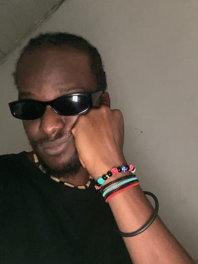 A medium shot, self-taken photograph features a Black male wearing sunglasses and a black t-shirt. He is resting his head on his right hand, with his left hand holding his right cheek. The man has a goatee and wears a beaded necklace. On his right wrist, he has several beaded bracelets with dice-shaped beads. The background is a plain, gray wall. The lighting is dim, suggesting an indoor setting. There is no identifiable location in Rahwol Chom, Nigeria.