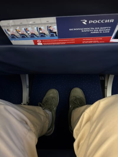 The image depicts a first-person, downward-facing perspective from a passenger seat inside an Airbus A319 aircraft, identified by the visible safety card. This scene is located at Емельяново (аэропорт), Russia.
In the foreground, the lower legs and feet of a person are visible. The individual is wearing light beige or khaki-colored trousers. Their feet are clad in olive green athletic shoes with matching laces, worn over white socks. The shoes feature a solid sole and a visible swoosh-like logo on the side. The feet are positioned on a dark blue carpeted floor, which exhibits a subtle pattern of light-colored speckles.
Directly ahead, the back of a dark blue airplane seat is visible, featuring a built-in seat-back pocket. A safety information card is inserted into this pocket. The card displays the airline name "РОССИЯ" (Rossiya) with its logo, and text in both Russian and English stating "БЕЗОПАСНОСТЬ НА БОРТУ" (Safety on Board) and "AIRBUS A319." The card also includes illustrative diagrams of passengers in safety-related positions within an aircraft cabin. A red band across the bottom of the card shows a "no smoking" symbol and corresponding text: "НЕ КУРИТЬ НА БОРТУ ВОЗДУШНОГО СУДНА" / "NO SMOKING AT ANYTIME," and instructions to "ВАМ НЕОБХОДИМО ВЫПОЛНЯТЬ УКАЗАНИЯ ЭКИПАЖА И СВЕТОВЫЕ ТАБЛО" / "FOLLOW DIRECTIONS OF THE CREW AND THE SIGNS." The environment is illuminated by artificial cabin lighting.