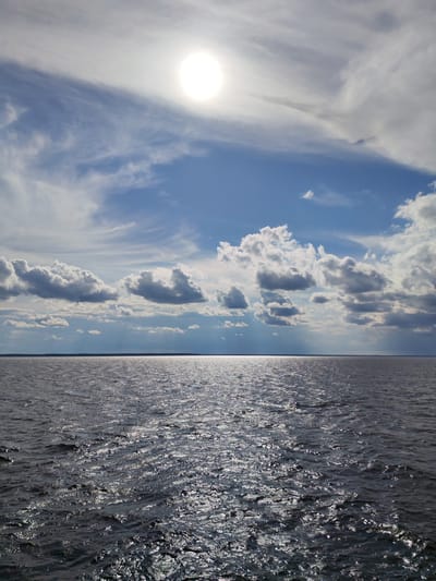 The image depicts a wide, calm body of water under a bright, partially cloudy sky. The sun is visible high in the sky, casting a bright glare on the water's surface, creating a shimmering effect. The water appears dark and slightly rippled, suggesting a gentle breeze. The horizon line is a thin strip separating the water from a distant, dark landmass. Fluffy white clouds are scattered across the blue sky, with some wispy clouds closer to the sun. The overall impression is one of a peaceful, expansive natural landscape. This scene is likely located in the vicinity of Myaksa, Russia, known for its proximity to Lake Beloye.