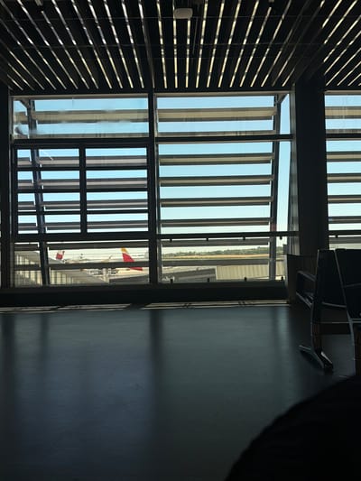 The image captures an interior view looking out through a large window with louvers. The scene appears to be an airport terminal or a similar facility with large windows overlooking an airfield. The window is divided into a grid of panes, with vertical louvers on the outside creating a striped effect.
Through the window, a section of an airplane is visible, painted white with a yellow and red tail. The airplane is parked at a gate or on the tarmac. In the distance, there are buildings and greenery, suggesting an airport environment.
The interior side of the window is dark, with what appear to be chairs or seating visible in the upper right portion of the frame. The overall lighting suggests it is daytime, possibly late afternoon or early morning given the angle of the light casting shadows. The weather appears to be clear.
The image is taken from a high vantage point, looking down and outwards. The composition emphasizes the contrast between the dark interior and the bright exterior, and the repetitive patterns of the louvers. There is no visible text in the image.
