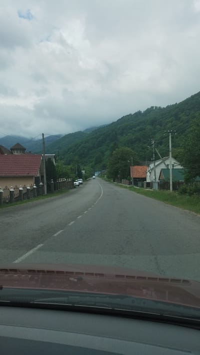 Captured from inside a vehicle, the image presents a road winding through the cityscape of Rakhiv, Ukraine. A mountain covered with lush, green forest dominates the left side of the frame, partially obscured by a cloudy sky. The road itself is gray asphalt with a white dividing line. Buildings with reddish-brown roofs are interspersed among the trees. The dashboard of the car is visible on the right side.