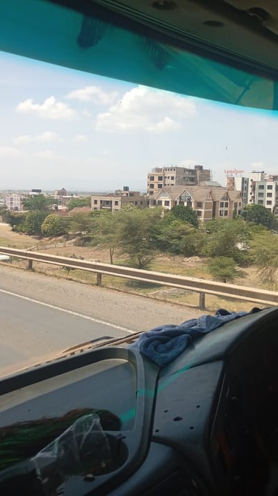 The image depicts an exterior view of Viwandani ward, Kenya, observed from the interior of a vehicle. The foreground shows the vehicle's dark grey dashboard, where a crumpled dark blue towel rests on the right side. On the left, within a dashboard recess, a clear plastic bag containing dark, possibly green and brown, fibrous material is visible. The top portion of the frame is obscured by a translucent green-blue tint of the windshield.
Beyond the vehicle, a two-lane asphalt road with a white solid edge line and a broken white center line runs horizontally. A light brown metal guardrail separates the road from a lower-lying area of dry, reddish-brown earth sparsely covered with small, thorny trees and green shrubs.
In the mid-to-far background, a cluster of buildings forms a cityscape. A prominent multi-story structure with a distinctive brown pitched roof, gables, and tan walls is identifiable as the "ANTIQUE SANTI HOTEL" by a sign on its facade. To its left and behind, several multi-story buildings in various stages of construction are visible, some with exposed concrete frames and rebar. To the right of the hotel, additional multi-story buildings, including one with a "Lemayan" sign on its side, contribute to the urban landscape. Green trees are interspersed among the structures and surrounding terrain. The sky is light blue with scattered white cumulus clouds, indicating sunny weather. No individuals are visible.