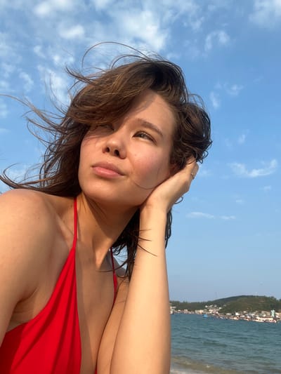 The image captures a young woman taking a selfie outdoors, likely by the sea. She is depicted from the chest up, with her head tilted slightly upwards. She wears a red bikini top and has dark, slightly wet hair that appears wind-swept. Her left hand is raised, gently touching her hair near her ear, revealing a small ring on her finger. Her eyes are partially closed or looking down, and she has a relaxed, subtle smile, conveying a sense of contentment.
The background features a bright blue sky with a few wispy white clouds. To the right, a body of water with small waves is visible, and beyond it, a distant coastline with green hills and scattered buildings. The bright, natural sunlight suggests the time of day is late morning or early afternoon, with clear and sunny weather. The scene clearly indicates a coastal or beach setting, consistent with the provided location context of Nam Natural Heritage Area, Trang, Vietnam. No text is visible in the image.
