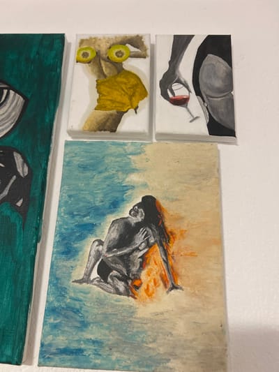 The scene is an interior view within Mosomishte, Bulgaria, displaying a collection of unframed painted canvases mounted on a plain white wall. A large, vertically oriented rectangular canvas is partially visible on the far left, featuring an abstract design with a stylized eye in black and white on a dark teal background. Above the central lower canvas, two smaller, vertically oriented rectangular canvases are arranged horizontally. The left of these depicts a stylized, light-brown colored nude torso with two yellow lemon slices positioned over the breasts and a crumpled yellow object obscuring the genital area. The right smaller canvas displays a dark grey hand holding a stemmed glass containing red liquid, positioned next to a rounded grey form, possibly a buttocks or breast, on a white background. Below these, a larger, vertically oriented rectangular canvas dominates the lower-center, illustrating two abstract, grey-toned human figures in an intimate embrace. The background of this painting transitions from shades of blue and white in the upper portion to intense orange and yellow, suggestive of fire or warmth, in the lower portion around the figures. The overall lighting is consistent, indicating an indoor setting.
