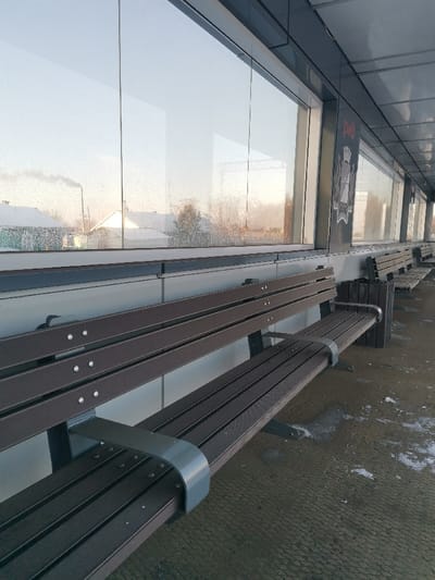 The image presents an unoccupied railway station platform or waiting area in Озеро-Карачи, Russia, under what appears to be winter or early spring conditions. The foreground and midground feature a series of dark brown, slatted benches with grey metallic support frames, extending linearly to the right. The ground is a dark, textured surface, likely concrete or asphalt, with distinct patches of snow and ice. A dark, slatted wooden trash bin is positioned next to the rightmost visible bench. In the background, a continuous glass wall, showing some frosting or dirt, separates the interior space from an exterior environment. Through this glass, snow-covered rooftops of residential structures are visible against a bright, hazy sky. A tall industrial chimney on the left emits a plume of white smoke. Bare trees and overhead railway infrastructure are also discernible beyond the glass. Above the glass wall, a dark grey ceiling or overhang extends, and a vertical dark panel displays the red "РЖД" (Russian Railways) logo along with an associated emblem. No individuals or specific activities are observed.