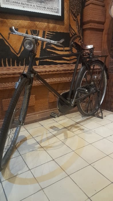 The image displays a vintage-style black bicycle positioned indoors in Glantengan, Indonesia. The bicycle stands upright on its kickstand on a light-colored tiled floor with dark grout lines, casting prominent shadows from a light source to the left. Key features of the bicycle include chrome drop handlebars, a circular headlight mounted on the front, full fenders covering both wheels, and a luggage rack at the rear. The background consists of a wooden wall adorned with intricate relief carvings or patterns in varying shades of brown. A framed picture, partially visible above the bicycle, contains text identifying it as "KELUARGA BAPAK H. A. SHOCHIB MABRURI". No individuals are present in the scene.