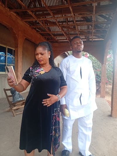 Two individuals are positioned outdoors in Ugwolawo, Nigeria, under an open-air structure featuring reddish-brown pillars, beams, and a corrugated metal roof.

On the left, a female subject, appearing middle-aged, stands holding a gold-toned smartphone with a textured case in her raised right hand. She wears a black velvet midi-dress with short sleeves, a V-neckline, and a multi-colored floral pattern on the upper left chest and a matching velvet panel with frog closures along a slit on the lower right side. Her left hand rests on her abdomen. She has dark hair styled in braids, gold hoop earrings, and multiple white beaded bracelets on her left wrist.

To her right, a male subject, appearing young adult, stands with his hands clasped in front of him, holding a rolled-up yellow object in his right hand. He is dressed in a white traditional two-piece outfit consisting of a long-sleeved tunic with black vertical detailing and matching trousers. He wears black slide sandals with a "TF" logo.

In the background, to the left of the female, a light-colored building with a single window is visible. A folded wooden chair sits behind her. Further back, on the right, green foliage with intermittent pink blossoms is present. The ground is unpaved and light brown.