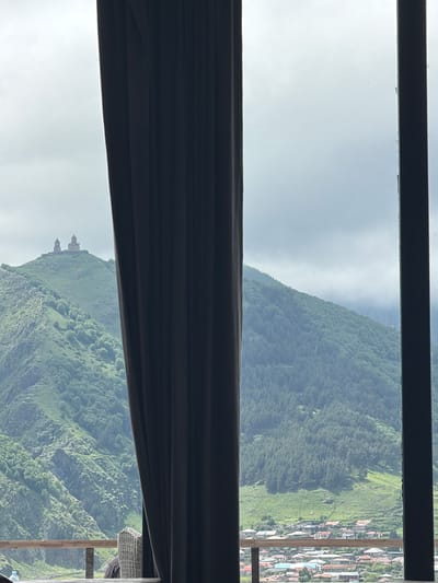 The image depicts a view from a window looking out over the city of Stepantsminda, Georgia. A dark curtain hangs on the left side of the frame, partially obscuring the scene. In the background, a prominent mountain rises, topped by the Gergeti Trinity Church, a historic landmark. The slopes of the mountain are covered in lush green vegetation, with scattered trees and rocky outcrops. Below the mountain, a portion of Stepantsminda is visible, characterized by a cluster of houses with terracotta roofs. The sky is overcast, with soft, diffused light. In the foreground, a portion of a wooden railing and a wicker chair can be seen, suggesting an outdoor seating area or balcony. A water bottle is also visible on the railing.