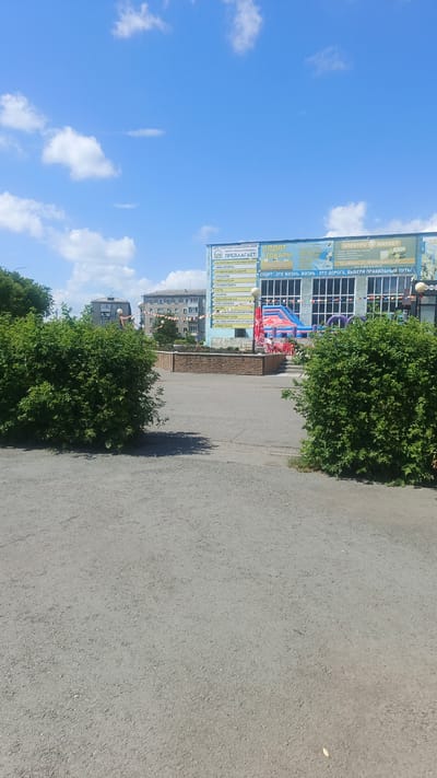 This is a photograph taken outdoors on a sunny day with a bright blue sky and scattered white clouds. The foreground is dominated by a paved area and two large, dense green bushes, one on the left and one on the right, casting shadows on the ground.

In the midground, there is a low, circular stone-faced wall, possibly part of a planter or seating area, with some red chairs visible behind it. Beyond the wall, the facade of a building is prominent. The building has large windows and a prominent sign with Cyrillic text.

The sign is a prominent feature, advertising "SPORT GOODS" and listing various services and activities available. Text visible on the sign includes "ПРЕДЛАГАЕТ:" (offers), "БАСТРЯН МАЛАТИ БОЛЬШАЯ ЗАРНА" (possibly related to gymnastics or sports), "АКВААЭРОБИКА" (aqua aerobics), "ГРУДНИЧКОВОЕ ПЛАВАНИЕ" (baby swimming), "ГИДРОМАССАЖ" (hydro-massage), "СОЛЯНАЯ ПЕЩЕРА" (salt cave), "САУНА" (sauna), "Солярий" (solarium), "ТРЕНАЖЕРНЫЙ ЗАЛ" (gym), "ФИТНЕС" (fitness), and "ВОСТОЧНЫЕ ТАНЦЫ" (oriental dances). It also includes an address: "ТД «Маяк», ул. Маяковского, 48" (Marketplace "Mayak", Mayakovsky St., 48) and a phone number. A slogan reads "СПОРТ - ЭТО ЖИЗНЬ. ЖИЗНЬ - ЭТО ДОРОГА. ВЫБЕРИ ПРАВИЛЬНЫЙ ПУТЬ!" (Sport is life. Life is a road. Choose the right path!). Another part of the sign mentions "ЭЛЕКТРО МАРКЕТ" (Electro Market).

In the background, several multi-story buildings are visible, suggesting an urban or town setting. Some are residential buildings, while the main building with the sign appears to be a commercial establishment or community center. There are also strings of festive decorations hanging between buildings, indicating a possible event or celebration. The overall impression is a bright, somewhat commercialized area in a Russian town. The time of day appears to be late morning or early afternoon, given the strong sunlight and shadows.