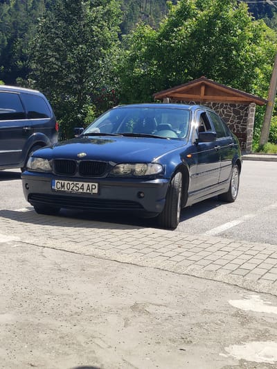 In the city of Krushev Dol, Bulgaria, a dark blue BMW sedan is parked on an asphalt surface adjacent to a cobblestone pathway. The car is facing towards the viewer, angled slightly to the right. Its license plate, which reads "CM 0254 AP" with "BG" in a blue section, is clearly visible. The car's headlights are on, and its windows are slightly open. To the left of the BMW, a dark-colored minivan is partially visible. In the background, a small, wooden-roofed structure with stone walls stands against a backdrop of lush green trees and a forested hillside. The lighting suggests it is daytime with bright sunlight, casting shadows on the ground.