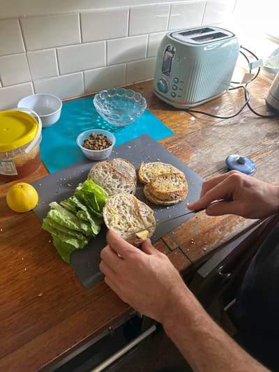 The image depicts a kitchen scene where someone is preparing sandwiches. A person's hands are visible, one holding a knife and spreading butter on a slice of bread. Several other slices of bread, some with butter already spread, are laid out on a dark gray cutting board. A bunch of fresh green lettuce leaves is positioned to the left of the bread. Also on the cutting board are some small dark brown pellets, possibly kibble or seeds, in a small clear plastic container. To the left of the cutting board, a bright yellow-lidded plastic container holds a golden, viscous substance, likely honey. A round yellow lemon sits next to the honey container. In the background, there are various kitchen items, including a light blue retro-style toaster, a white bowl, a small clear decorative bowl, and a tiled backsplash. The scene is set on a wooden countertop. There is no indication of the location being in the city of Thora, Australia.