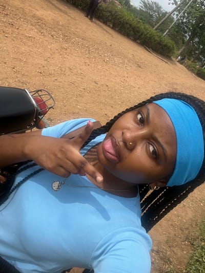 The image displays a young Black female positioned in the foreground, capturing a selfie in Gbongan, Nigeria. She wears a light blue short-sleeved top and a matching light blue fabric headband over dark braided hair. Her right hand is raised, forming a "shaka" gesture with extended thumb and pinky, and her fingernails are painted light pink. Her tongue is slightly extended from her mouth. She has a gold stud piercing on her right nostril and small gold hoop earrings. A thin gold chain necklace with a silver, embellished pendant, possibly resembling Hello Kitty, hangs around her neck. The subject appears to be seated on or leaning against a dark-colored motorcycle, with its rear seat and a metal rack partially visible in the lower-left. The background consists of an unpaved reddish-brown dirt surface scattered with small stones. Beyond this, a line of green shrubs and distant trees are visible. A blurred figure in dark attire stands in the upper-mid-background. The scene is outdoors under daylight.