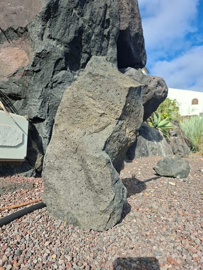 The image depicts an outdoor scene with volcanic rocks in Santiago del Teide, Spain. A large, dark gray, rough-textured volcanic rock formation dominates the left side of the frame. In the foreground, a medium-sized, lighter gray, porous volcanic rock stands upright. The ground is covered with a layer of small, reddish-brown and gray gravel. A black tube or hose runs horizontally across the lower left corner of the image, partially obscured by gravel. In the background, a white building with a arched window and part of a red-tiled roof is visible. Lush green vegetation, including a succulent plant and tall grass, grows behind the rocks. The sky above is blue with scattered white clouds. The lighting suggests a bright, sunny day.