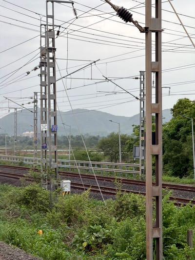 The image depicts a view of railway tracks and overhead electrical infrastructure in Vasai-Virar, India, under an overcast sky. In the foreground, a tall, light-brown metal pole with a square cross-section stands prominently, supporting numerous wires. The pole is part of a larger network of similar poles and complex electrical lines that extend into the distance.
To the left of the main pole, another similar pole is visible, also supporting electrical infrastructure. Attached to this pole are two rectangular signs. The upper sign features a white background with black markings, appearing to be a diagram or symbol. Below it, a blue sign with white lettering, possibly indicating a route or designation, is visible. Further into the background, more poles and electrical lines are present, crisscrossing over the railway tracks.
The railway tracks themselves are a focal point, with ballast visible beneath the rails. The tracks appear to be part of a multiple-track railway system, with at least two sets of parallel tracks discernible.
The surrounding environment is lush and green, with dense vegetation and trees bordering the railway line. In the middle ground, a white electrical box with yellow and black striped markings is situated near the tracks. The background is dominated by rolling hills or mountains covered in vegetation, fading into the hazy sky. Streetlights are also visible along the railway line in the middle distance.
No people are visible in the image. The scene is static and focused on the infrastructure and natural surroundings of the railway line. The overall impression is one of a functional, operational railway corridor in a semi-urban or rural setting within Vasai-Virar.