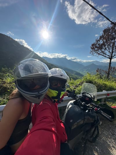 Two individuals in motorcycle helmets are depicted in a selfie-style shot, taken on a sunny day in a mountainous region identified as Tú Lệ, Vietnam.

The subject on the left wears a light gray full-face motorcycle helmet with its clear visor partially raised, revealing dark sunglasses underneath. Their left shoulder and a dark tank top are visible. The subject on the right, partially obscured by the left individual and positioned slightly behind, wears a white and yellow full-face helmet, also with dark sunglasses visible. They are attired in a red long-sleeved jacket.

A dark gray or black adventure-style motorcycle is parked beside them on a paved road. Visible components include its handlebars, a clear front windshield, fuel tank, and engine crash bars. The motorcycle is positioned next to a silver metal guardrail.

The environment features a clear blue sky with scattered white clouds. A prominent sunburst emanates from the upper-left sky, casting strong light across the scene. The background consists of multiple layers of green, tree-covered mountains, receding into a hazy blue distance. Green terraced fields are discernible in the valleys between the distant mountains. Lush green vegetation and shrubs line the immediate roadside embankment. On the far right, a solitary, defoliated tree with dark, intricate branches stands against the sky.