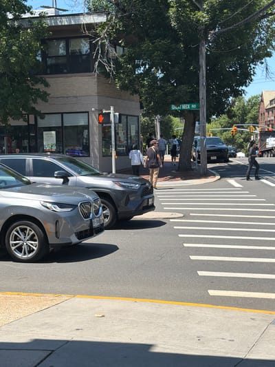 The image depicts a street scene in the Village of Great Neck Plaza, United States of America, on a sunny day. In the foreground, two cars are visible: a silver BMW SUV on the left and a gray Toyota RAV4 on the right. Both vehicles are positioned on the road, facing towards the right of the frame. A crosswalk with white stripes is prominent in the middle ground, indicating a pedestrian crossing.

Several pedestrians are present in the scene. On the sidewalk to the left, a person wearing a white hoodie and dark pants is standing. Further down the sidewalk, a group of people are walking. Across the street, near the intersection, a person wearing a gray long-sleeved shirt and dark pants is crossing the street. Another person in dark clothing is walking on the sidewalk on the right side of the street.

A traffic light with a red hand signal is visible on a pole to the left, suggesting that pedestrians have the right of way at this moment. A green street sign reading "GREAT NECK RD" is affixed to a wooden utility pole in the middle of the frame. In the background, a dark-colored Dodge Challenger is parked on the street. A large tree with dense foliage partially obscures the view of the buildings in the background. A multi-story building with brick and beige siding is situated on the corner, and a portion of another brick building is visible on the far right. The environment appears to be an urban or suburban commercial area with trees lining the street and sidewalks.