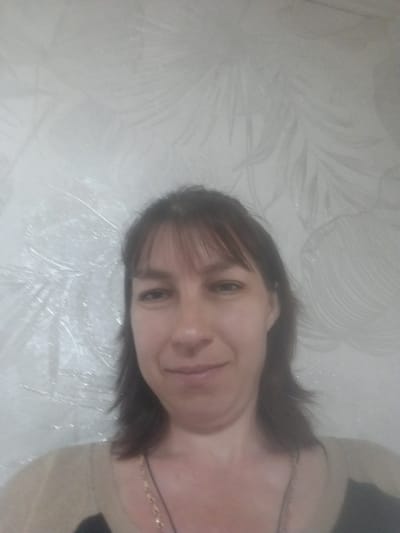 Here is a description of the image:
A woman with brown hair and bangs is seen in a close-up, slightly angled shot. She is wearing a light-colored sweater with a black top underneath. A thin necklace is visible around her neck. The background appears to be a light-colored wall with a subtle, embossed pattern of abstract shapes or leaves. The overall image is somewhat blurry, possibly due to low lighting or camera focus. The location is unspecified in Бородулиха ауылдық округі, Kazakhstan.