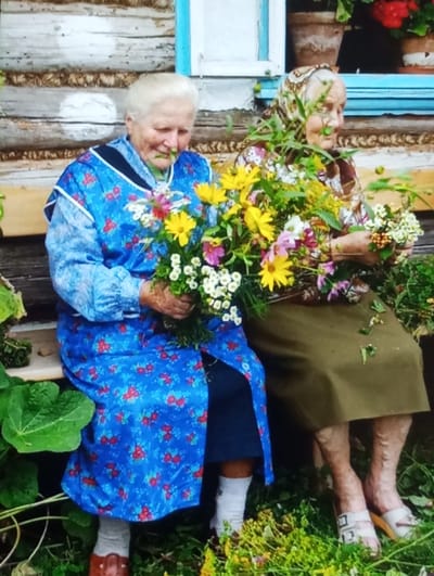 Two elderly women are seated on a wooden bench outdoors in Wojnowice, Poland. The woman on the left has white hair and wears a blue long-sleeved dress with a red floral pattern over a dark skirt, white socks, and brown closed-toe shoes. She holds a large bouquet of assorted wildflowers, including prominent yellow, white, and pink blooms, along with green foliage. The woman on the right wears a brown and white patterned headscarf, a floral print blouse, and an olive green skirt, with white open-toed sandals. She holds smaller bunches of white flowers and green foliage in her hands. Both individuals are looking down at the flowers they are holding. The background consists of a log-constructed building wall with a light blue-trimmed window, upon which potted plants with red flowers are visible. Green vegetation, including broad leaves, is present on the ground around the bench.