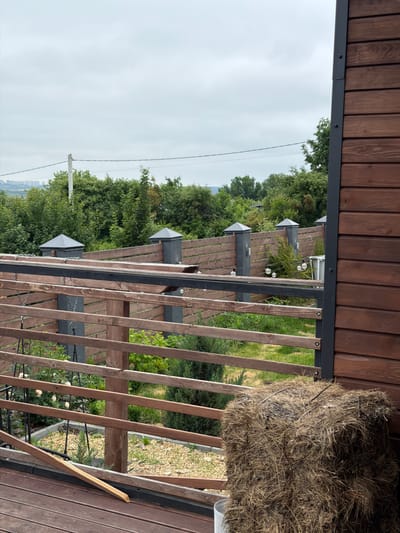 The image shows a view from a wooden deck looking outwards towards a backyard area. In the foreground on the right, a large bale of hay is positioned next to a wooden wall of a building. The deck has wooden planks and a wooden railing with horizontal slats. Beyond the railing, there is a grassy area with some small evergreen trees and white roses. A wooden fence with dark grey posts topped with pyramid-shaped caps runs along the perimeter of the backyard. In the distance, a line of trees and a hazy cityscape are visible under an overcast sky. The overall scene suggests a residential area in or near the city of Semiluki, Russia.
