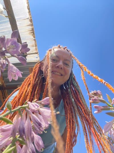 In the village of Tatarskaya Kargala, Russia, a woman with bright orange dreadlocks is captured in a low-angle, upward-facing shot. She is smiling and looking towards the camera, partially obscured by clusters of delicate, pale purple flowers in the foreground. Behind her, the clear blue sky is visible, with the edge of a weathered, translucent corrugated roof structure on the left side of the frame. The woman is wearing a light blue t-shirt. The scene appears to be outdoors, possibly in a garden or near a greenhouse.