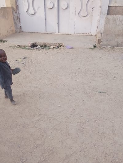 A young child stands barefoot in a sandy area in front of a white metal gate. The child is wearing a dark outfit and holds something in one hand. A small, circular pile of debris and litter sits on the ground near the gate, with a glimpse of greenery. The background shows portions of a beige building. The area appears arid.