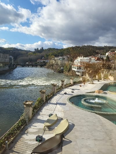 The image depicts a riverfront scene in São Pedro do Sul, Portugal, under a partially cloudy sky. The Vouga River occupies the left side of the frame, flowing from the background towards the foreground, with visible rapids and sun reflections on its surface. A multi-arched stone bridge crosses the river in the mid-distance.
On the right side of the image, a paved promenade extends parallel to the river. Three empty, curved chaise lounges are positioned on this promenade: two light-colored and one dark-colored. Adjacent to the promenade, a multi-tiered circular water feature, possibly a thermal pool or fountain, displays turquoise water with an active central jet. A stone balustrade with dark metal railings and green foliage separates the promenade from the river.
Buildings line both banks of the river beyond the bridge. On the left bank, modern structures are visible. On the right bank, a cluster of white and light-colored buildings with multiple stories and balconies extends along the river and up the hillside. The background features densely tree-covered hills under a sky with both blue patches and large cumulus clouds. No individuals are present in the scene.