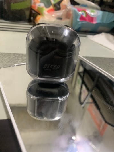 The image shows a black wireless earbud case with a clear lid, reflecting on a mirrored surface. The brand name "BISEN" is visible on the side of the case. In the background, out of focus, are some colorful items, possibly packaging or other consumer goods. The environment appears to be indoors, with a striped surface beneath the case and a lighter colored surface below that. There are no people or specific landmarks of Angeles City, Philippines, visible in the image.