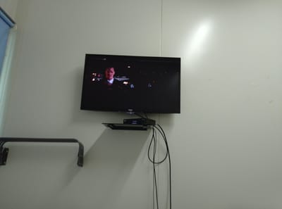The image shows a flat-screen television mounted on a white wall. The television is displaying a movie scene featuring a man in a suit. Below the television, a black shelf is attached to the wall, holding a black electronic device with a blue digital display showing "0046". Several black cables hang from the device. To the left of the television, a metal bracket is mounted on the wall, casting a shadow. The wall appears to be indoors and has a textured surface. A light fixture is visible on the upper right side of the wall, casting a bright reflection. The overall impression is a domestic or office setting. The location is specified as Қопа ауылдық округі, Kazakhstan, implying this is a scene within that region.