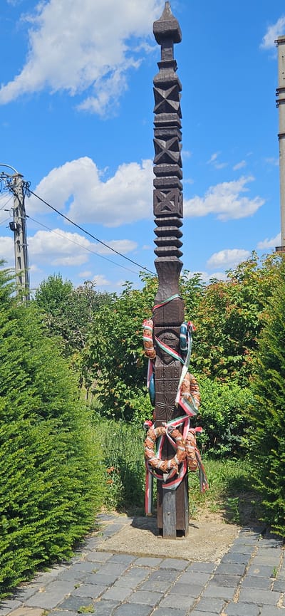 A carved wooden pole stands upright in a paved area surrounded by green bushes and trees under a blue sky with white clouds. The pole is dark brown and features intricate geometric carvings throughout its length. It is decorated with two wreath-like ornaments and several ribbons in red, white, and green, suggesting a connection to Hungary. The lower section of the pole shows carved details that appear to be numbers "1848" and "1998", possibly indicating a significant historical event or anniversary. The ground in front of the pole is paved with dark grey square stones, transitioning to a concrete base around the pole's base, which is then surrounded by gravel. Electrical poles and wires are visible to the left of the main subject, and a tall, unfinished concrete structure is partially visible to the right. The scene appears to be an outdoor memorial or landmark.