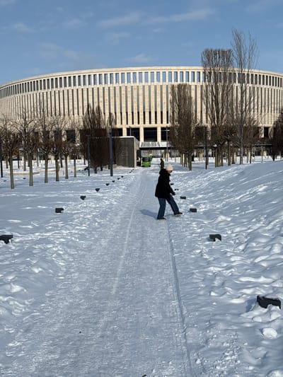 The image displays an outdoor winter scene in the Plodorodny microdistrict of Krasnodar, Russia, featuring Krasnodar Stadium under a clear blue sky. The stadium, a large, circular, multi-story structure, dominates the background with its light-colored, ribbed facade composed of numerous vertical rectangular window sections.
In the mid-ground, a solitary individual is seen walking on a cleared path of compacted snow and ice that leads towards the stadium. This person, with their back partially to the camera, wears a dark jacket, blue jeans, light-colored footwear, and a white beanie. Their right leg is lifted and extended, indicating a careful step on the potentially slippery surface.
The ground on either side of the path is covered with undisturbed, undulating snow. Small, dark, rectangular blocks are partially visible embedded in the snow, arranged in rows parallel to the path. Numerous bare deciduous trees are symmetrically planted in rows across the mid-ground, flanking the path and the stadium. In the far background, near a stadium entrance, a small green utility vehicle is visible alongside several indistinct figures. The overall environment is brightly lit by sunlight, consistent with a clear winter day.