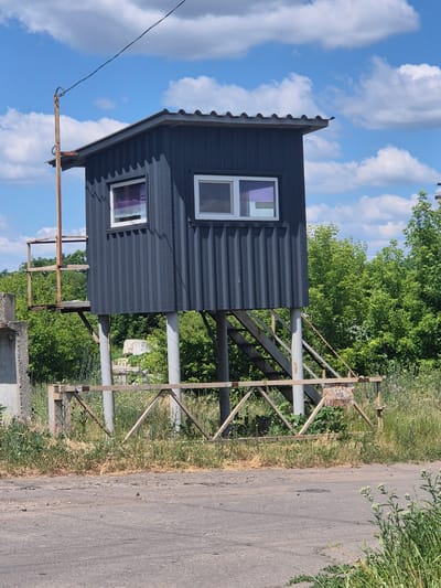 A dark gray, corrugated metal guard booth stands elevated on four cylindrical concrete pillars in Sozonivka, Ukraine. The booth has a sloped roof and two visible windows with white frames. Through one window, a purple curtain is visible. The other window has a sign that reads "ОХОРОНА" (Ohorona), meaning "Guard" or "Security" in Ukrainian. A metal staircase with railings leads up to the booth. A rusty metal structure, possibly a former guard platform or a support for an antenna, is attached to the left side of the booth. In front of the booth, a simple wooden barrier with metal supports crosses a gravel and dirt path. The area is surrounded by lush green trees and foliage. The sky above is blue with scattered white clouds. The overall scene appears to be in a rural or semi-rural setting.