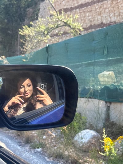The image, taken in the Municipal Unit of Sfakiotes, Greece, shows a view through a car's side mirror. The reflection in the mirror features a woman holding a smartphone, with her hand gesturing towards the phone's camera. The background outside the car window appears to be a landscape with rocks, foliage, and structures covered with green mesh. A glimpse of a building or wall is visible beyond the mesh. Sunlight illuminates parts of the scene.