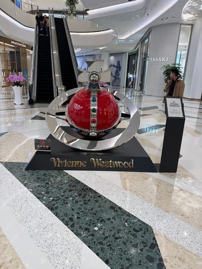 Here is a description of the image:

The image captures a Vivienne Westwood display in what appears to be a high-end shopping mall, likely located in Jianye District, China, based on the Simplified Chinese signage. The display features a large, red orb with silver rings and decorative jewels, all presented on a black stand with the brand name "Vivienne Westwood" in gold lettering. A sign reading "请勿踩踏 Do not stampede" is placed near the base of the display.

In the background, an escalator is visible, with a few people riding it. Other storefronts are noticeable, including a "TASAKI" store and a "SHIATZY CHEN" store. The mall has modern architecture, characterized by curved white walls, marble floors with geometric patterns, and recessed lighting. A man in a tan jacket can be seen looking at his phone near the TASAKI store.