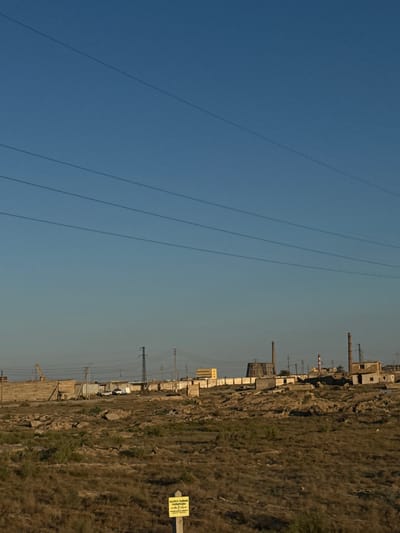 The image shows a landscape with a clear blue sky on the left and a dry, grassy terrain on the right. Several power lines stretch across the sky. In the distance, along the horizon where the sky meets the terrain, there is an industrial area with various structures, including chimneys and buildings, and numerous electrical pylons and power lines. The terrain appears arid and rocky with sparse vegetation.

In the right portion of the image, a yellow warning sign is visible, mounted on a pole. The sign has text written on it, appearing to be in a Cyrillic-based script. The sun is shining, suggesting it is daytime, likely late afternoon or early morning given the angle of the light. The scene depicts an industrial area adjacent to a natural, undeveloped landscape, suggesting a remote or semi-industrialized location. No people are visible in the image. The overall mood is utilitarian and somewhat desolate due to the industrial elements and dry terrain.