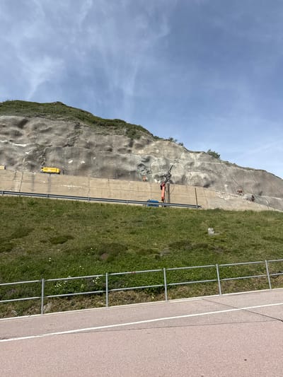 In Airolo, Switzerland, on a clear day with scattered clouds, a steep, rocky mountainside is covered with a protective mesh net. A concrete barrier runs horizontally across the lower portion of the slope, supported by a guardrail. To the left of the slope, a yellow generator sits on the concrete barrier. Further to the right on the barrier, a person in an orange vest operates a mechanical arm, with a blue platform visible below. Several other figures, some in orange vests, are scattered on the slope, seemingly engaged in work. A grassy embankment slopes down from the barrier, bordered by a metal fence and a paved path with a white line.