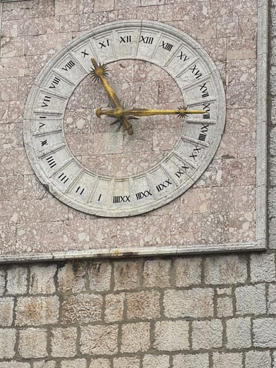 This is a close-up, eye-level, medium shot of a large, ornate clock face mounted on a stone wall in the city of Krk, Croatia. The clock face is circular and made of a light-colored material, possibly stone or marble, with Roman numerals indicating the hours. The numerals range from I to XXIV, suggesting a 24-hour clock. The clock has two golden hands; the hour hand is shorter and thicker, while the minute hand is longer and thinner, both featuring decorative tips. The clock is set against a wall constructed of rectangular stone blocks of varying sizes, with a reddish-pink marbled pattern visible behind the clock and a rougher, lighter-colored stone below. The clock is positioned on a building, as indicated by the architectural style of the surrounding stonework. The image captures the detail of the clock's Roman numerals and the texture of the stone.
