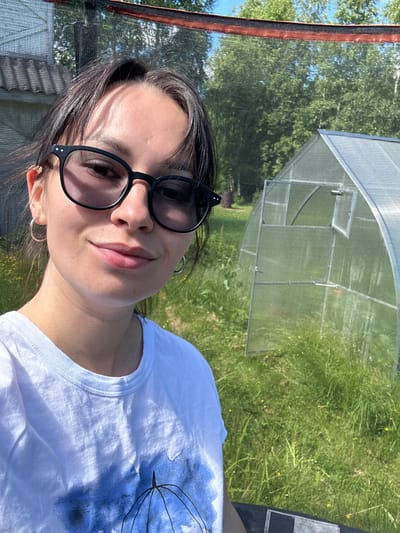 The image is a selfie taken outdoors, likely in the city of Novozhilovo, Russia, on a sunny day. A young woman with dark hair and glasses is in the foreground, smiling at the camera. She is wearing a white t-shirt with a blue abstract design. Behind her, to the right, is a small greenhouse with a transparent cover. The background consists of lush green grass, some scattered yellow wildflowers, and trees. A portion of a building with a tiled roof is visible to the left, and a black net structure with a red trim is present at the top of the frame, possibly part of a trampoline or a protective enclosure. The overall impression is a casual outdoor scene in a rural or suburban setting.