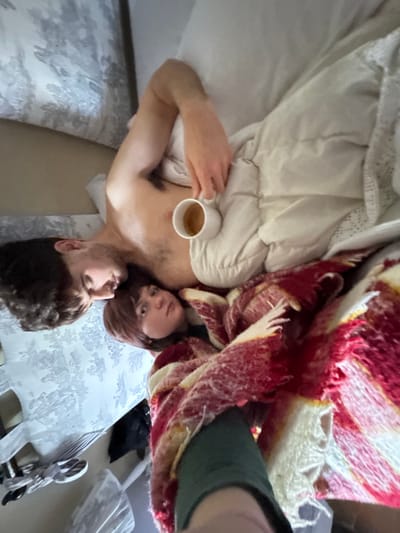 Two individuals, one male and one female, are observed lying on a bed in a residence located in Montepulciano, Italy. The male, positioned on the left side of the frame, is bare-chested with dark hair and a visible nose stud. He holds a white ceramic mug containing a light brown liquid in his right hand, which rests near his chest. He is partially covered by a light-colored duvet and is looking towards the female. The female, positioned to the male's right and slightly lower, has reddish-brown hair with bangs and is looking directly at the camera. Both individuals are partially covered by a textured red, white, and yellow patterned blanket. A limb, likely an arm or leg of a third individual, covered in dark green fabric, is visible in the lower foreground, indicating the perspective of the camera holder. The bed features patterned pillowcases with a light floral design. Miscellaneous items, including what appears to be footwear, are partially visible on a surface to the left of the bed.
