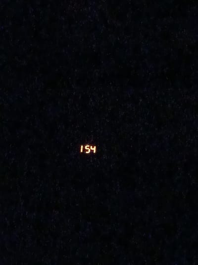 This is a close-up, dark, and grainy photograph of a digital display showing the number "154" in an orange glow. The background is almost entirely black, with some subtle speckling of what might be dust, debris, or a textured surface. The lighting is very dim, with the only significant light source being the illuminated digits. The image does not contain any people or reveal any specific setting beyond the immediate digital display. There are no weather cues, location cues, or emotional indicators present. The number "154" is the sole piece of visible text. The provided location context of Ekaterinovka, Russia, does not provide any visual cues within the image itself.
