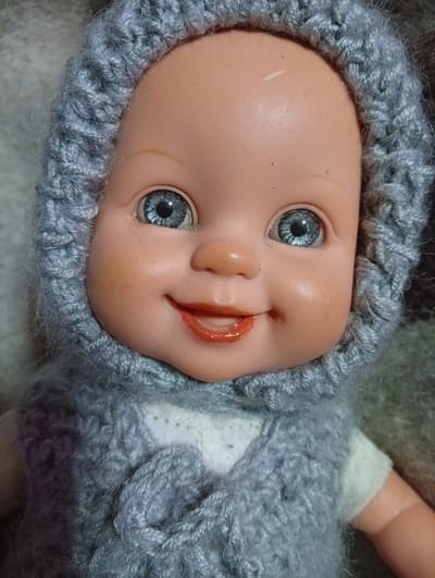 This is a close-up studio portrait of a doll wearing a grey knitted outfit, which includes a bonnet and a vest. The doll has blue eyes, a small nose, and painted red lips that are slightly parted in a smile. The background is a textured, neutral-colored fabric. There is no indication of location within the city of Ostrov-Zalit, Russia.