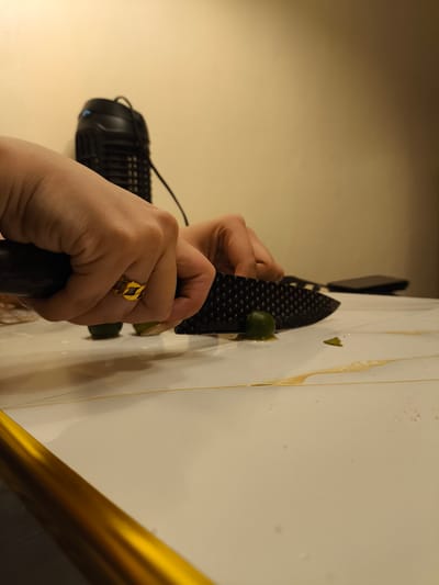 The image shows a close-up of a person's hands as they cut small, green fruits, likely calamansi, with a black knife. The fruits are on a white countertop with marble-like veining and some spilled juice. The person is wearing a gold ring on their left hand. In the background, a black object, possibly a mosquito trap or a fan, is visible against a beige wall. The lighting suggests it might be indoors, possibly at night or in a dimly lit room. The overall scene depicts a food preparation activity.