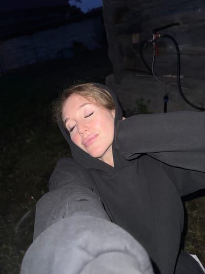 A young woman with closed eyes and light-colored hair is seen in a close-up, selfie-style shot. She is wearing a black hooded sweatshirt and appears to be outdoors at night, possibly in Maksimovo, Russia, given the provided context. The background is dark and indistinct, showing some grassy terrain and what appears to be a rustic structure with pipes. The lighting suggests a low-light environment, with the woman's face illuminated by what could be artificial light or the camera's flash. Her pose, with hands near her head and eyes closed, conveys a sense of relaxation or sleep.