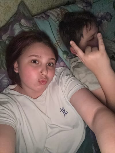 This media file captures two individuals indoors, likely in a bedroom setting, in Lyman, Ukraine. The lighting is dim, suggesting it is evening or night.

In the foreground, a young female with reddish-brown hair is lying down, looking directly at the camera. She has noticeable eyebrow and nose piercings. Her lips are pursed in a playful "kissing face" expression, and her eyes are wide open, conveying a lighthearted or affectionate mood. She is wearing a white t-shirt, which features a dark, stylized logo or symbol on its left chest area.

Partially visible in the background, behind and above the female, is another individual with dark hair. This person is also lying down, with their face partially obscured by a hand from the foreground individual, which has two fingers extended playfully. The person in the background is wearing a light-colored t-shirt.

Both individuals are resting on bedding or pillows adorned with a patterned design in shades of green, purple, and blue. The overall scene suggests a casual and intimate moment, possibly a shared selfie or photo.